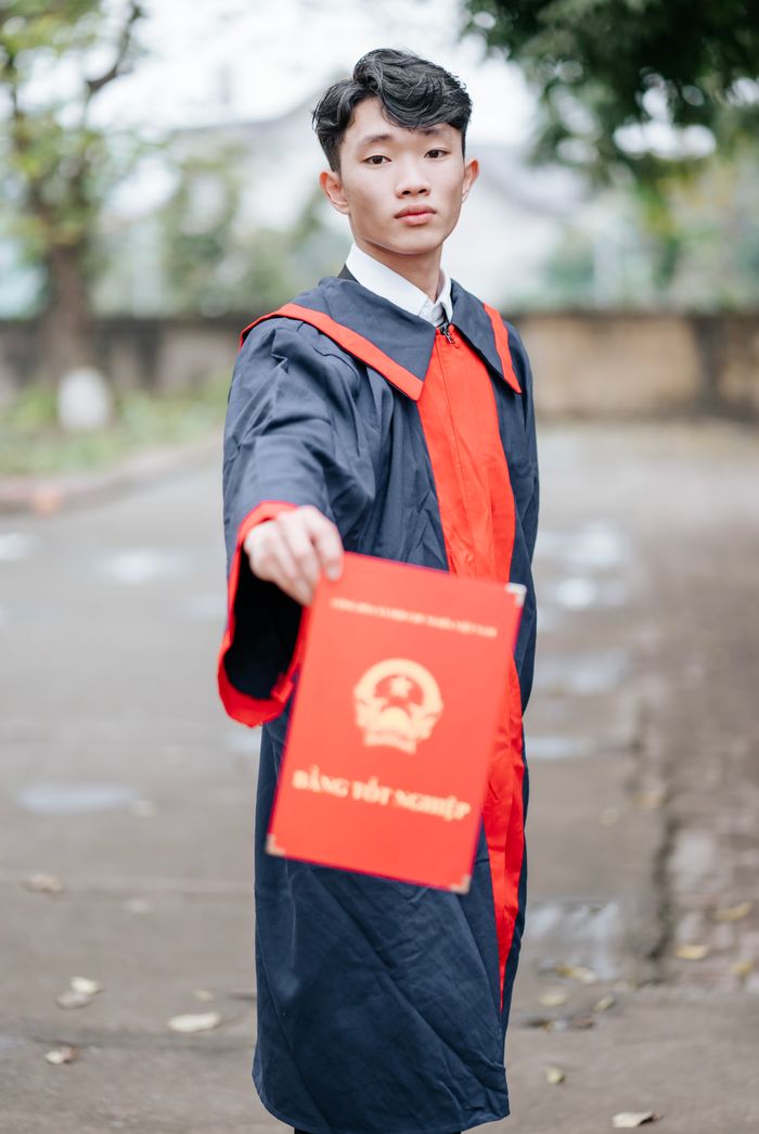 Home Graduate in cap and gown holding a red certificate outdoors, celebrating achievement.