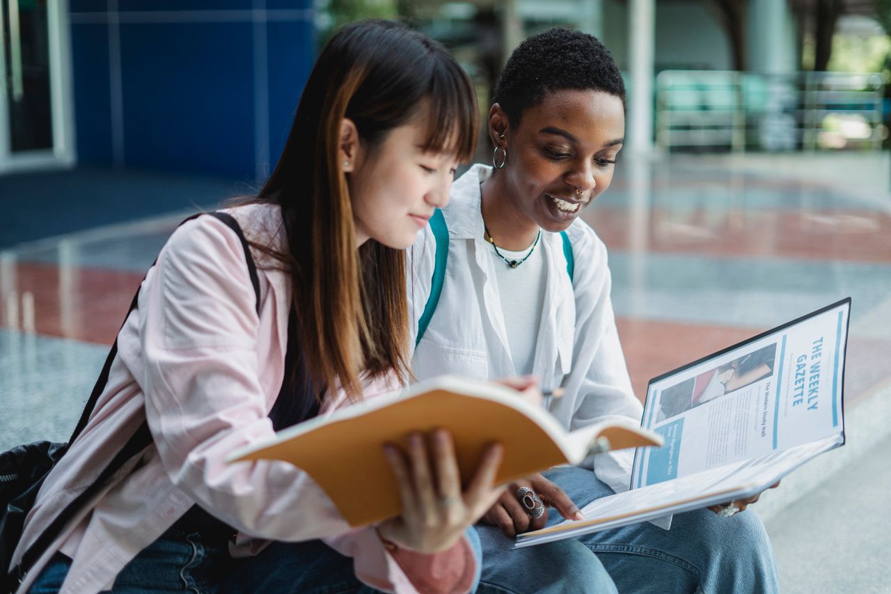 Home Young content multiethnic best female friends speaking while watching textbook on street stairs in city