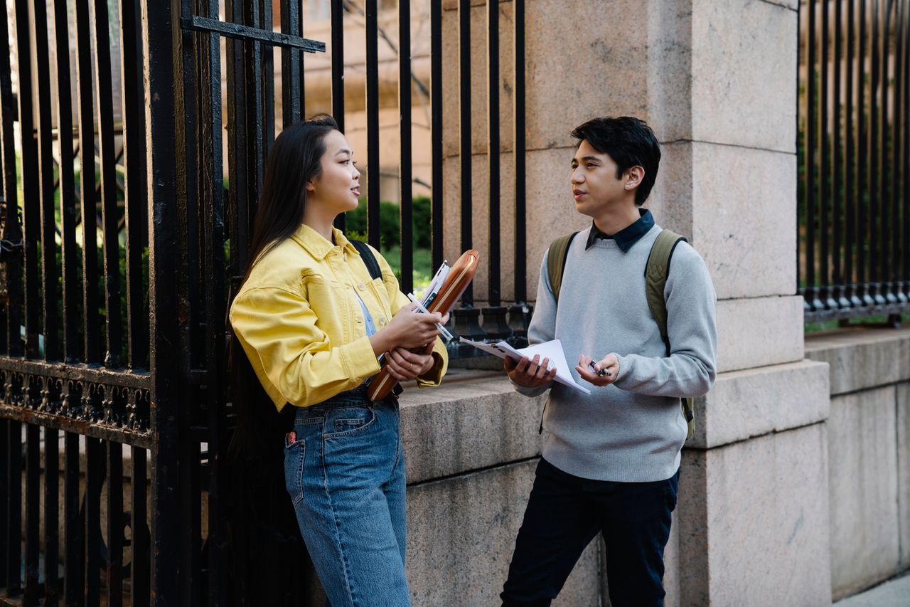 Home Two young adults engaged in conversation near a university gate.