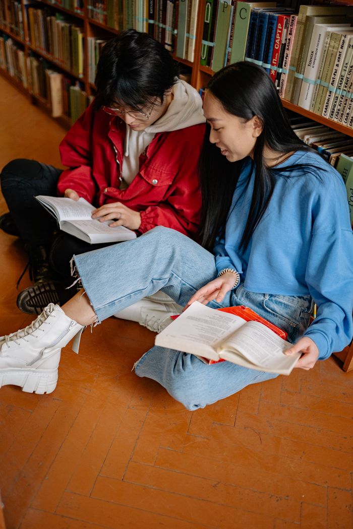Two students sitting on library floor reading books, enjoying a study session together.